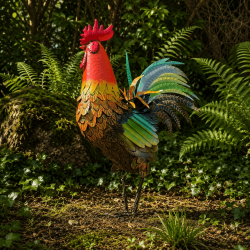 Großer Metallhahn mit bunten Flügeln zwischen grünen Gartenpflanzen