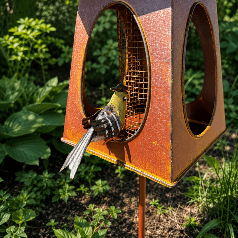 Farbenfroher Metallvogel vor dem Gitterbehälter der Vogelfutterstelle